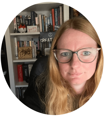 A woman who hates having a photo taken, with long red hair, wearing glasses, sitting in front of a bookcase.